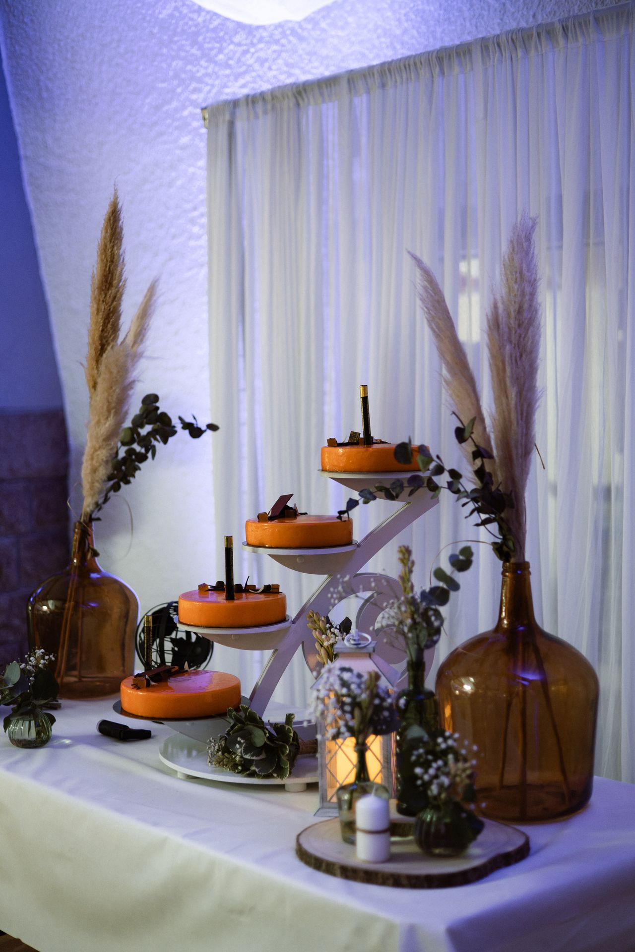 Mariage Salle de la Goubaudière espace gateaux table blanche grand vase transparent de couleur et pampas rondin en bois et feuillage lanternes rideaux blanc