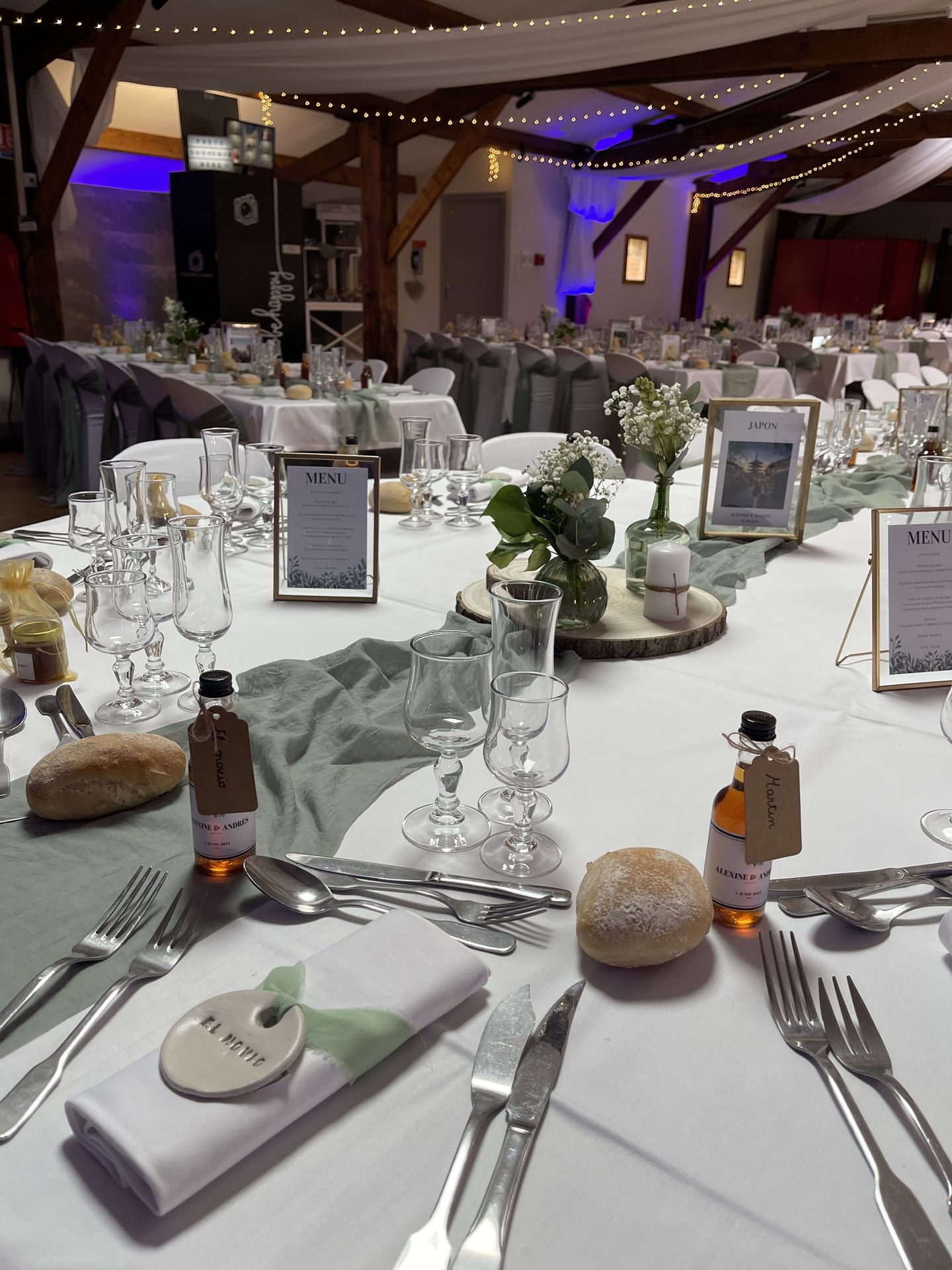 Mariage Salle de la Goubaudière salle intérieure tables blanches chemin de table vert sauge fiole en verre cadeaux invités centre de tabler rondin en bois composition florales et feuillage serviette noeuds vert et nom invités