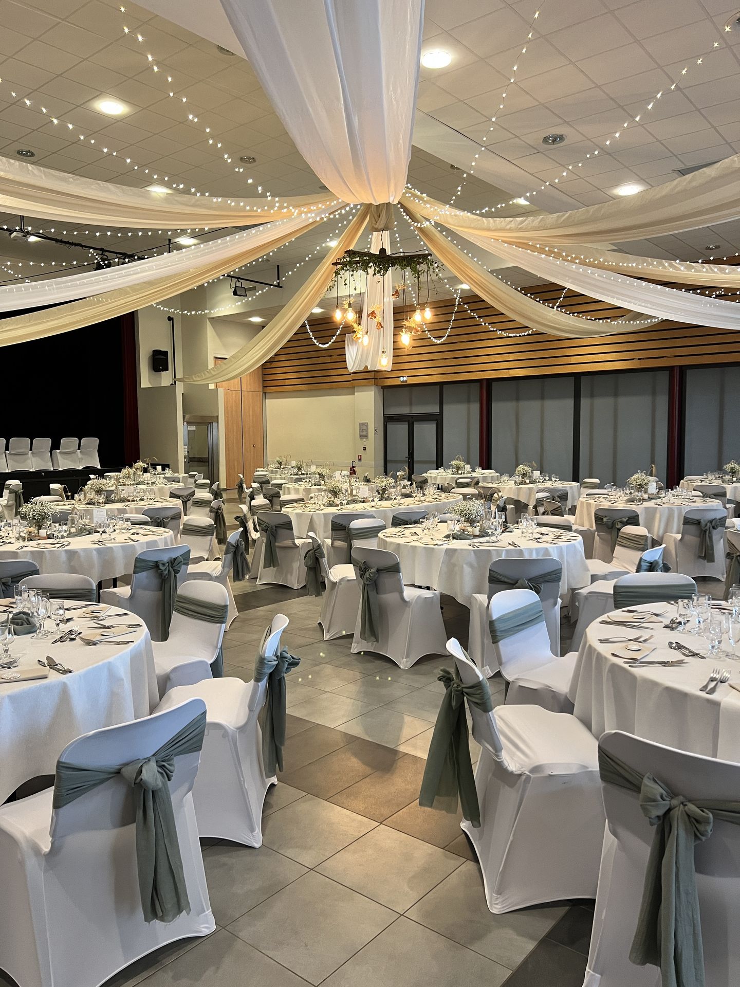 Mariage Espace Galerne plafond tentures blanches et sables structures bois feuillage et ampoules guinguette et vase pampas table blanche et noeud vert sauge