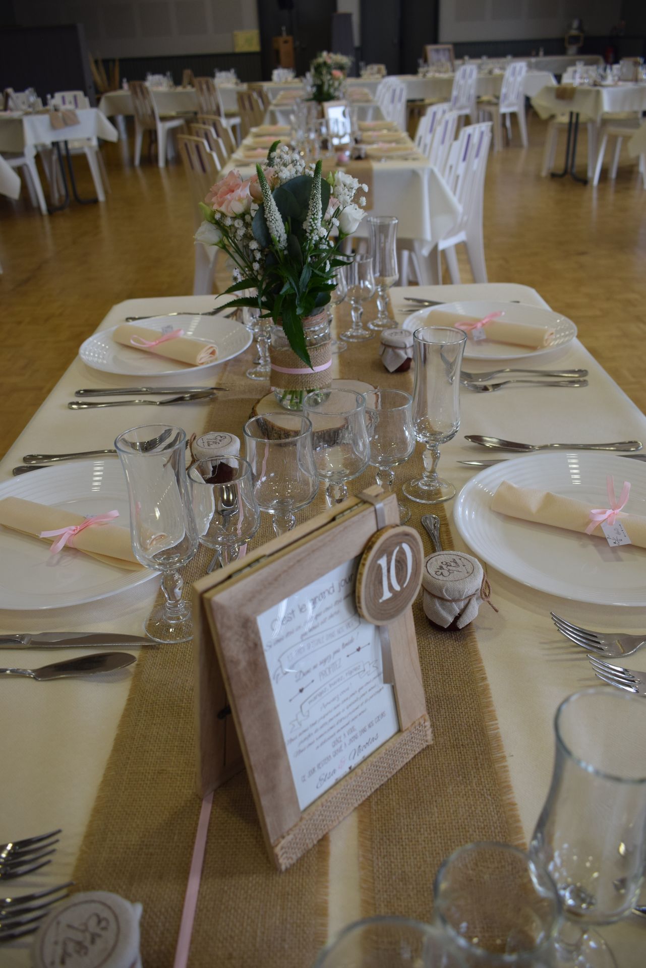 Mariage Salle L'Arc en Ciel table blanche chemin de table sable cadre en bois numéro de table rondin en bois vase de fleurs serviette beige et noeud rose
