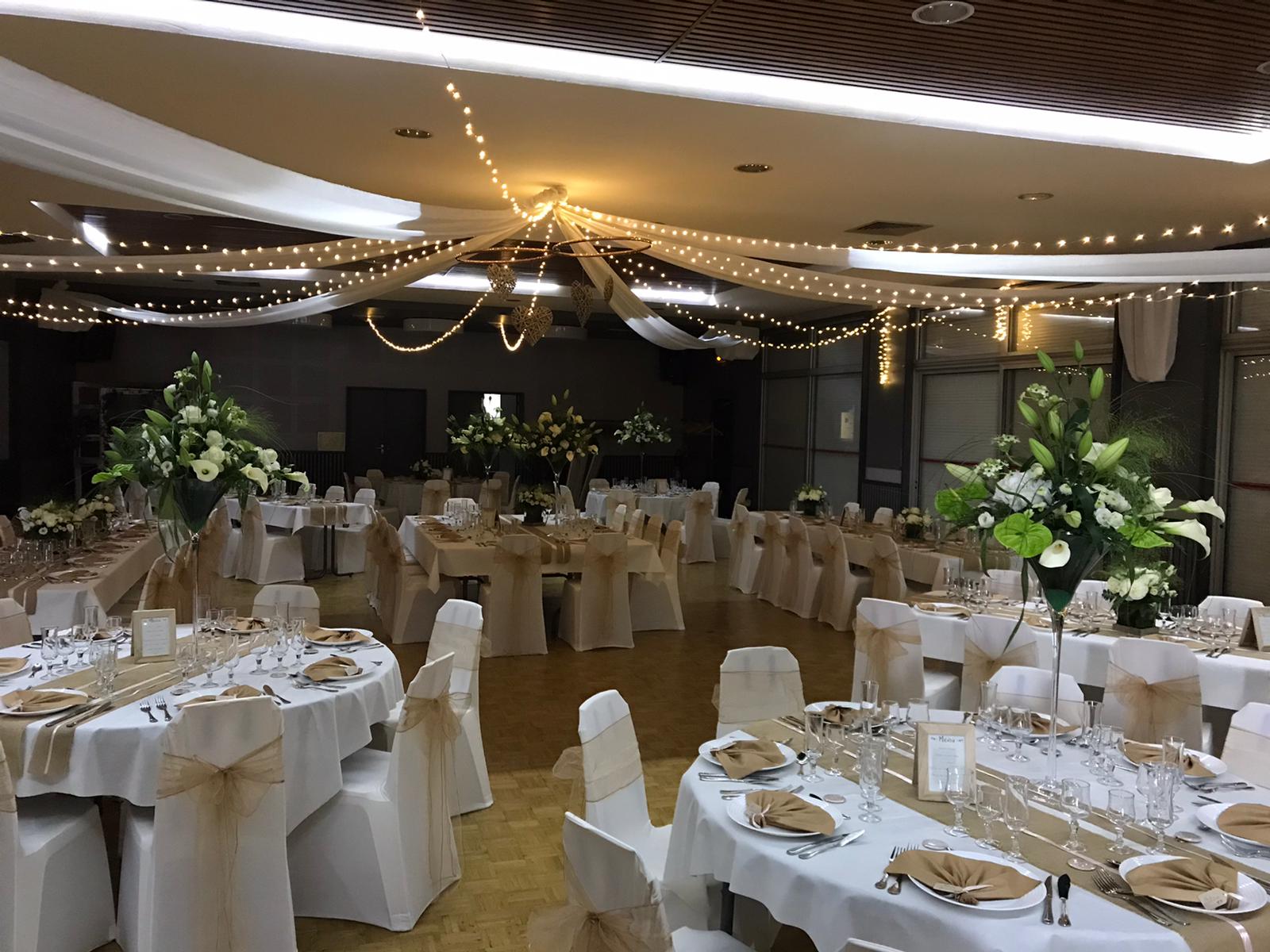 Mariage Salle L'Arc en Ciel plafond tentures blanches et leds coeurs en maille naturel table blanche chemin de table sable et noeud de chaise sable composition florale
