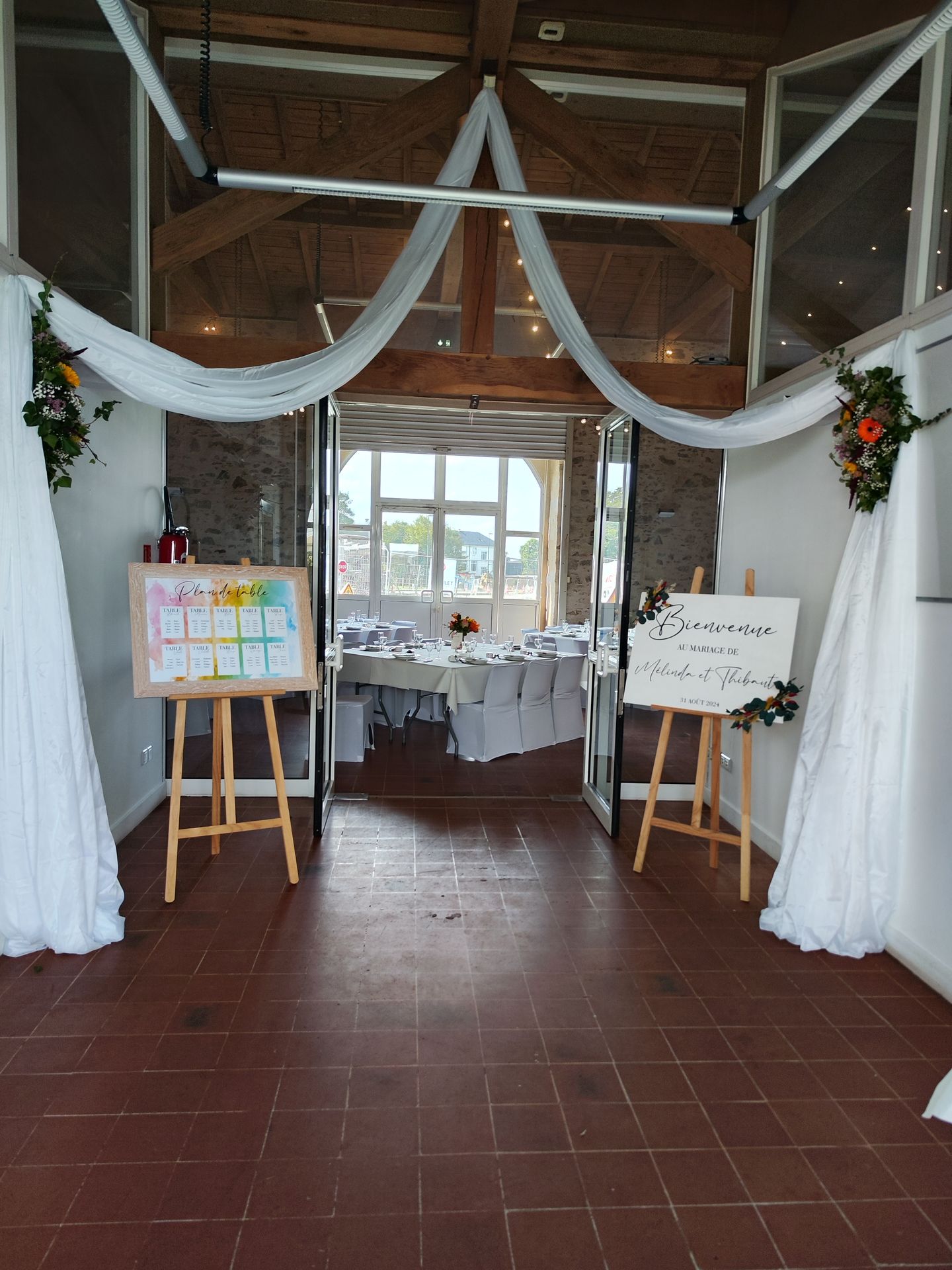 Mariage Salle de l'Etenduère entrée salle tentures blanches composition florale panneau de bienvenue blanc fleurs sèche plan de table arc en ciel panneau en bois